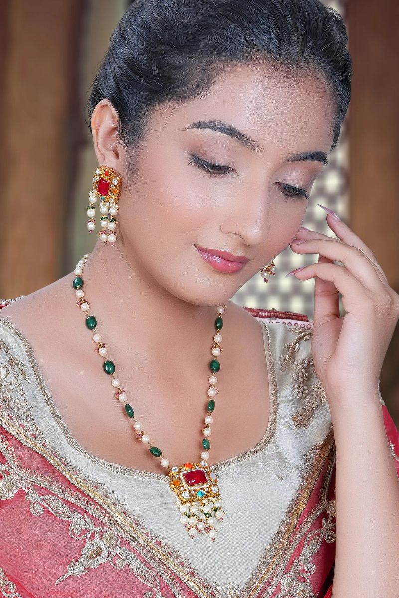 Woman wearing a traditional necklace with red and green stones, posing with hand near face.